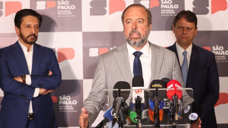 Esta é uma fotografia tirada em um ambiente interno, durante o que parece ser uma coletiva de imprensa ou anúncio oficial, focada em três figuras políticas masculinas em primeiro plano, com microfones e um painel de fundo. O primeiro, falando para microfones, é Alexandre Silveira. Atrás dele, aparecem Ricardo Nunes e Tarcísio de Freitas.