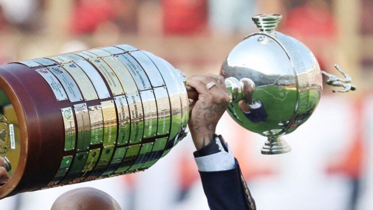 A foto mostra um registro em close-up e em ângulo lateral da Taça Libertadores da América sendo erguida por um jogador. O troféu está na horizontal, ocupando grande parte do quadro.