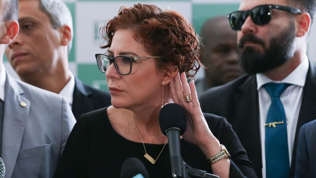 A imagem é um close-up que foca na deputada federal Carla Zambelli em um ambiente formal, possivelmente uma coletiva de imprensa ou evento político, com outras pessoas em segundo plano.