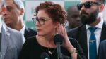 A imagem é um close-up que foca na deputada federal Carla Zambelli em um ambiente formal, possivelmente uma coletiva de imprensa ou evento político, com outras pessoas em segundo plano.