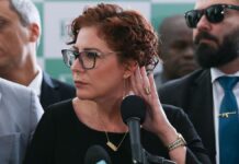 A imagem é um close-up que foca na deputada federal Carla Zambelli em um ambiente formal, possivelmente uma coletiva de imprensa ou evento político, com outras pessoas em segundo plano.