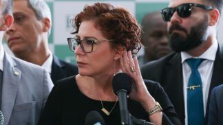 A imagem é um close-up que foca na deputada federal Carla Zambelli em um ambiente formal, possivelmente uma coletiva de imprensa ou evento político, com outras pessoas em segundo plano.