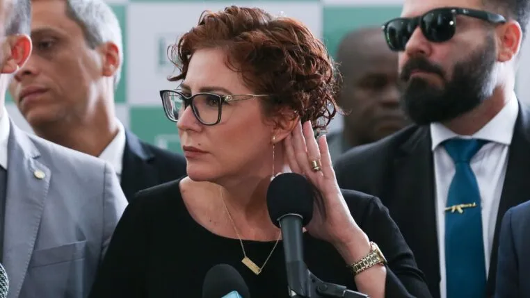 A imagem é um close-up que foca na deputada federal Carla Zambelli em um ambiente formal, possivelmente uma coletiva de imprensa ou evento político, com outras pessoas em segundo plano.