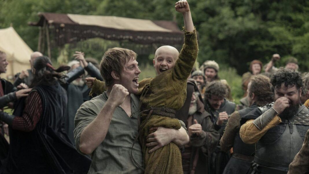 Homem levanta uma criança careca nos braços enquanto ambos comemoram com punhos erguidos; ao redor, um grupo de pessoas vestidas com trajes medievais celebra em um acampamento ao ar livre, com tendas e vegetação ao fundo.