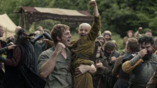 Homem levanta uma criança careca nos braços enquanto ambos comemoram com punhos erguidos; ao redor, um grupo de pessoas vestidas com trajes medievais celebra em um acampamento ao ar livre, com tendas e vegetação ao fundo.