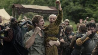 Homem levanta uma criança careca nos braços enquanto ambos comemoram com punhos erguidos; ao redor, um grupo de pessoas vestidas com trajes medievais celebra em um acampamento ao ar livre, com tendas e vegetação ao fundo.