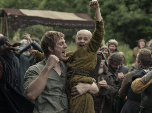 Homem levanta uma criança careca nos braços enquanto ambos comemoram com punhos erguidos; ao redor, um grupo de pessoas vestidas com trajes medievais celebra em um acampamento ao ar livre, com tendas e vegetação ao fundo.