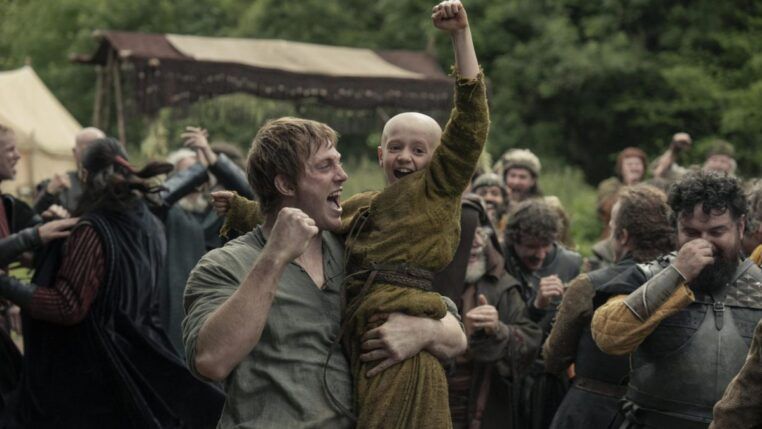 Homem levanta uma criança careca nos braços enquanto ambos comemoram com punhos erguidos; ao redor, um grupo de pessoas vestidas com trajes medievais celebra em um acampamento ao ar livre, com tendas e vegetação ao fundo.