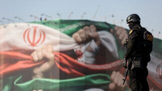 Um policial fortemente armado observa um grande mural ao ar livre que retrata a bandeira do Irã com punhos cerrados, simbolizando resistência; a cena ocorre em um espaço público sob céu claro.