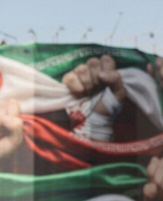 Um policial fortemente armado observa um grande mural ao ar livre que retrata a bandeira do Irã com punhos cerrados, simbolizando resistência; a cena ocorre em um espaço público sob céu claro.