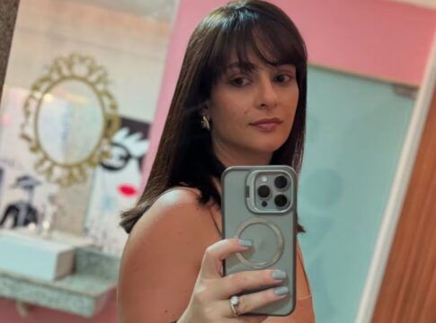 Mulher de cabelos castanhos, com franja, tira uma selfie em frente ao espelho usando um celular; ela veste roupa sem mangas e está em um ambiente interno com paredes em tons de rosa e azul e objetos decorativos ao fundo.