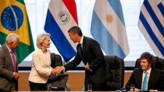 Autoridades participam de um encontro oficial: uma mulher de blazer claro cumprimenta um homem de terno escuro com um aperto de mãos, enquanto outros líderes observam sentados à mesa; ao fundo, bandeiras de países da América do Sul e do Brasil indicam um contexto diplomático ou internacional.