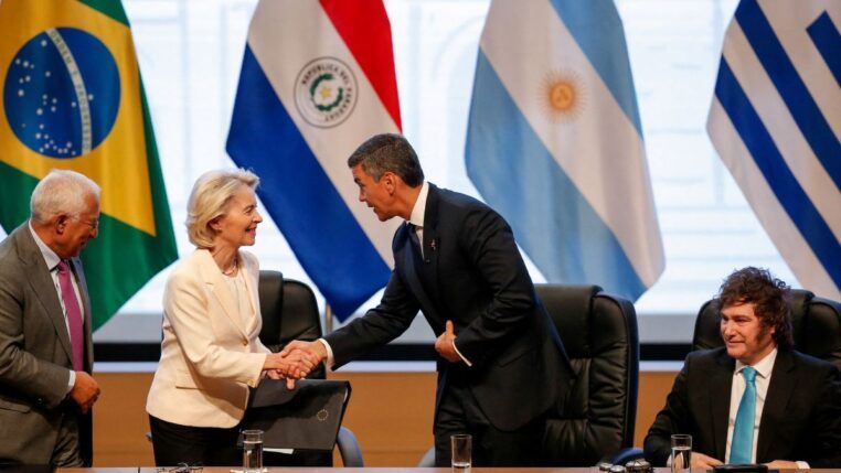 Autoridades participam de um encontro oficial: uma mulher de blazer claro cumprimenta um homem de terno escuro com um aperto de mãos, enquanto outros líderes observam sentados à mesa; ao fundo, bandeiras de países da América do Sul e do Brasil indicam um contexto diplomático ou internacional.