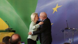 Uma mulher de cabelos loiros e um homem de barba branca se cumprimentam com um abraço em um palco oficial; ao fundo, aparecem elementos das bandeiras do Brasil e da União Europeia, além de púlpitos e microfones.