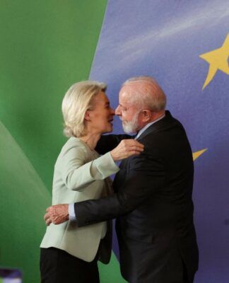 Uma mulher de cabelos loiros e um homem de barba branca se cumprimentam com um abraço em um palco oficial; ao fundo, aparecem elementos das bandeiras do Brasil e da União Europeia, além de púlpitos e microfones.