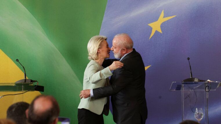 Uma mulher de cabelos loiros e um homem de barba branca se cumprimentam com um abraço em um palco oficial; ao fundo, aparecem elementos das bandeiras do Brasil e da União Europeia, além de púlpitos e microfones.