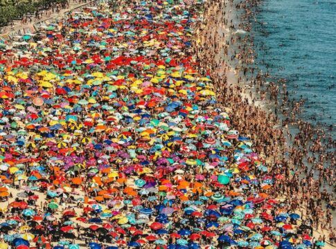 Vista aérea de uma praia lotada, com milhares de pessoas espalhadas pela areia e pelo mar, e uma grande concentração de guarda-sóis coloridos formando um mosaico vibrante ao longo da orla.