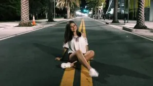 Foto de Maisa há dez ano sentada em uma rua com palmeiras na calçada