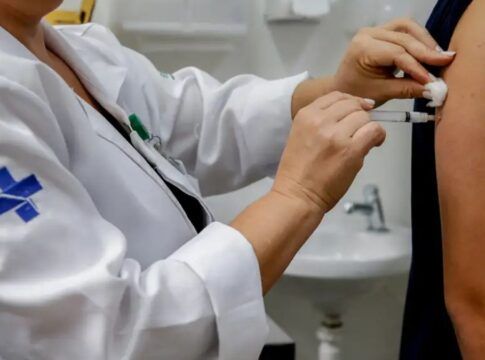 Enfermeira vestida com as roupas do SUS aplica uma vacina no braço de uma pessoa.