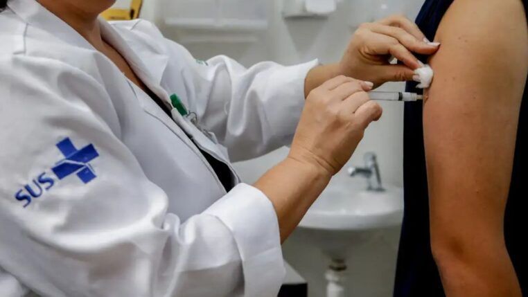 Enfermeira vestida com as roupas do SUS aplica uma vacina no braço de uma pessoa.