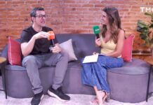 Um homem e uma mulher estão sentados em um sofá cinza curvo, participando de uma entrevista em um estúdio com parede de tijolos aparentes. O homem, à esquerda, usa óculos, camiseta preta e calça cinza, segurando um microfone laranja com o logotipo 