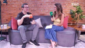 Um homem e uma mulher estão sentados em um sofá cinza curvo, participando de uma entrevista em um estúdio com parede de tijolos aparentes. O homem, à esquerda, usa óculos, camiseta preta e calça cinza, segurando um microfone laranja com o logotipo "TMC". A mulher, à direita, usa uma blusa amarela e calça jeans, segurando um microfone verde também com a marca "TMC". Ambos sorriem e parecem engajados em uma conversa descontraída.