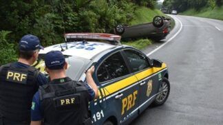 Em primeiro plano, dois policiais rodoviários federais de costas estão atrás de uma viatura da PRF estacionada na margem da rodovia. Eles observam um carro de passeio capotado às em curva sinuosa.