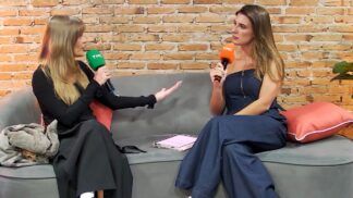 Duas mulheres estão sentadas em um sofá cinza, conversando e segurando microfones com a marca TMC — um microfone verde (à esquerda) e um laranja (à direita).