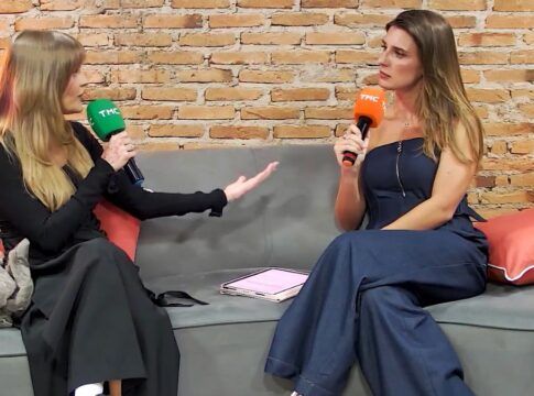 Duas mulheres estão sentadas em um sofá cinza, conversando e segurando microfones com a marca TMC — um microfone verde (à esquerda) e um laranja (à direita).