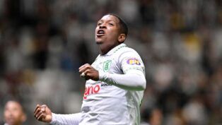 Um jogador de futebol veste o uniforme branco do América-MG e celebra com os punhos cerrados e o olhar voltado para cima. Ele utiliza uma camisa de mangas longas com detalhes verdes na gola e o escudo do clube no peito. Ao fundo, as arquibancadas do estádio estão lotadas, criando um cenário desfocado de torcedores. Outro jogador, também de branco, aparece parcialmente no canto inferior esquerdo da imagem.