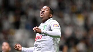 Um jogador de futebol veste o uniforme branco do América-MG e celebra com os punhos cerrados e o olhar voltado para cima. Ele utiliza uma camisa de mangas longas com detalhes verdes na gola e o escudo do clube no peito. Ao fundo, as arquibancadas do estádio estão lotadas, criando um cenário desfocado de torcedores. Outro jogador, também de branco, aparece parcialmente no canto inferior esquerdo da imagem.