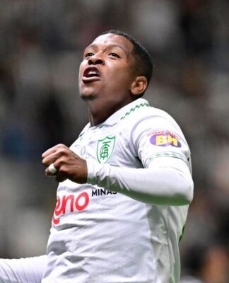 Um jogador de futebol veste o uniforme branco do América-MG e celebra com os punhos cerrados e o olhar voltado para cima. Ele utiliza uma camisa de mangas longas com detalhes verdes na gola e o escudo do clube no peito. Ao fundo, as arquibancadas do estádio estão lotadas, criando um cenário desfocado de torcedores. Outro jogador, também de branco, aparece parcialmente no canto inferior esquerdo da imagem.