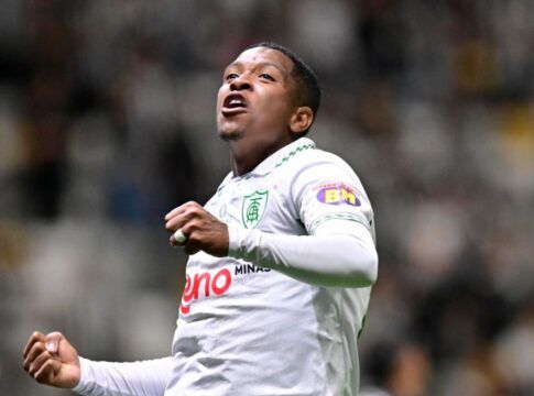 Um jogador de futebol veste o uniforme branco do América-MG e celebra com os punhos cerrados e o olhar voltado para cima. Ele utiliza uma camisa de mangas longas com detalhes verdes na gola e o escudo do clube no peito. Ao fundo, as arquibancadas do estádio estão lotadas, criando um cenário desfocado de torcedores. Outro jogador, também de branco, aparece parcialmente no canto inferior esquerdo da imagem.