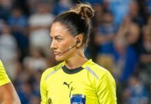 A árbitra de futebol Daiane Muniz é vista de perfil durante uma partida. Ela tem os cabelos castanhos presos em um coque alto e firme, e utiliza um sistema de comunicação (headset) no ouvido. Veste o uniforme oficial de arbitragem na cor amarelo vibrante com detalhes em preto. Sua expressão é séria e concentrada. Ao fundo, as arquibancadas do estádio aparecem desfocadas com torcedores vestindo azul.