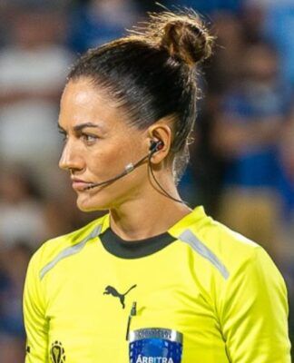 A árbitra de futebol Daiane Muniz é vista de perfil durante uma partida. Ela tem os cabelos castanhos presos em um coque alto e firme, e utiliza um sistema de comunicação (headset) no ouvido. Veste o uniforme oficial de arbitragem na cor amarelo vibrante com detalhes em preto. Sua expressão é séria e concentrada. Ao fundo, as arquibancadas do estádio aparecem desfocadas com torcedores vestindo azul.