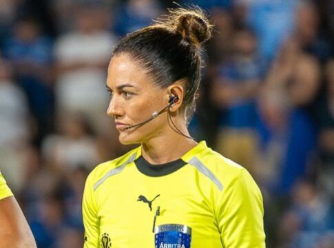 A árbitra de futebol Daiane Muniz é vista de perfil durante uma partida. Ela tem os cabelos castanhos presos em um coque alto e firme, e utiliza um sistema de comunicação (headset) no ouvido. Veste o uniforme oficial de arbitragem na cor amarelo vibrante com detalhes em preto. Sua expressão é séria e concentrada. Ao fundo, as arquibancadas do estádio aparecem desfocadas com torcedores vestindo azul.