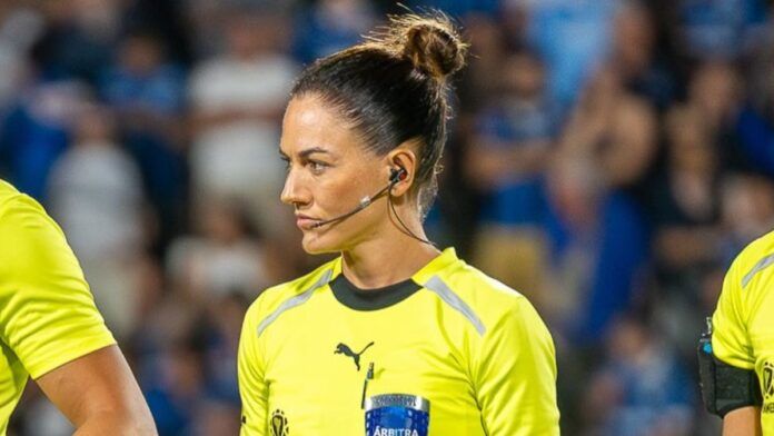 A árbitra de futebol Daiane Muniz é vista de perfil durante uma partida. Ela tem os cabelos castanhos presos em um coque alto e firme, e utiliza um sistema de comunicação (headset) no ouvido. Veste o uniforme oficial de arbitragem na cor amarelo vibrante com detalhes em preto. Sua expressão é séria e concentrada. Ao fundo, as arquibancadas do estádio aparecem desfocadas com torcedores vestindo azul.