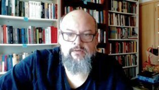 Um homem de pele clara, calvo e com barba grisalha cheia olha para a câmera. Ele usa óculos de armação escura e uma camiseta preta. Ao fundo, há prateleiras repletas de livros organizados, sugerindo um escritório ou biblioteca doméstica. A iluminação é frontal.