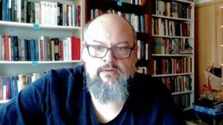 Um homem de pele clara, calvo e com barba grisalha cheia olha para a câmera. Ele usa óculos de armação escura e uma camiseta preta. Ao fundo, há prateleiras repletas de livros organizados, sugerindo um escritório ou biblioteca doméstica. A iluminação é frontal.