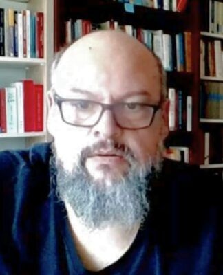 Um homem de pele clara, calvo e com barba grisalha cheia olha para a câmera. Ele usa óculos de armação escura e uma camiseta preta. Ao fundo, há prateleiras repletas de livros organizados, sugerindo um escritório ou biblioteca doméstica. A iluminação é frontal.