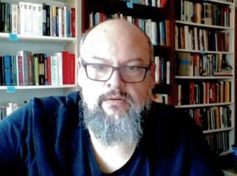 Um homem de pele clara, calvo e com barba grisalha cheia olha para a câmera. Ele usa óculos de armação escura e uma camiseta preta. Ao fundo, há prateleiras repletas de livros organizados, sugerindo um escritório ou biblioteca doméstica. A iluminação é frontal.