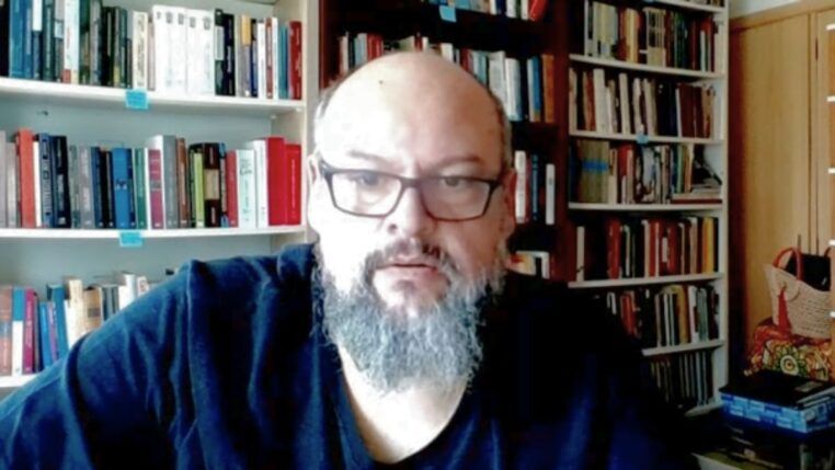 Um homem de pele clara, calvo e com barba grisalha cheia olha para a câmera. Ele usa óculos de armação escura e uma camiseta preta. Ao fundo, há prateleiras repletas de livros organizados, sugerindo um escritório ou biblioteca doméstica. A iluminação é frontal.