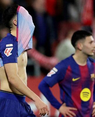 Dois jogadores de futebol em campo com uniformes azul e grená. O jogador em primeiro plano está de perfil, com a camisa puxada cobrindo o rosto, revelando o tronco. Ao fundo, outro jogador observa com as mãos na cintura. O clima sugere desânimo ou frustração.