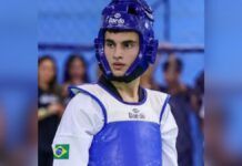 Um jovem atleta de taekwondo é visto do busto para cima, vestindo o equipamento de proteção oficial. Ele utiliza um capacete de proteção azul com a marca 