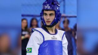 Um jovem atleta de taekwondo é visto do busto para cima, vestindo o equipamento de proteção oficial. Ele utiliza um capacete de proteção azul com a marca "Daedo" na testa e um protetor de tórax (dobok) também azul sobre uma vestimenta branca. No braço direito da vestimenta branca, há uma pequena bandeira do Brasil. O jovem tem pele clara e cabelos escuros, mantendo uma expressão séria e focada enquanto olha para o lado. O fundo da imagem está desfocado em tons de azul.