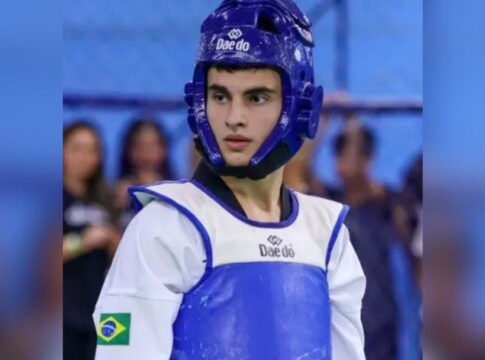 Um jovem atleta de taekwondo é visto do busto para cima, vestindo o equipamento de proteção oficial. Ele utiliza um capacete de proteção azul com a marca 