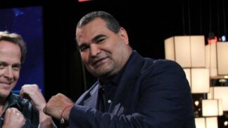 O ex-goleiro paraguaio José Luis Chilavert, um homem de pele clara e porte robusto, sorri levemente para a câmera. Ele veste um blazer azul-marinho sobre uma camisa social da mesma cor. No pulso direito, carrega uma pulseira de fio vermelho. À esquerda, parte do rosto de outro homem sorridente é visível, e o fundo escuro destaca luminárias cúbicas brancas à direita.