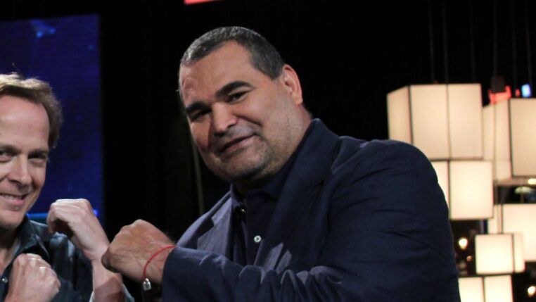 O ex-goleiro paraguaio José Luis Chilavert, um homem de pele clara e porte robusto, sorri levemente para a câmera. Ele veste um blazer azul-marinho sobre uma camisa social da mesma cor. No pulso direito, carrega uma pulseira de fio vermelho. À esquerda, parte do rosto de outro homem sorridente é visível, e o fundo escuro destaca luminárias cúbicas brancas à direita.