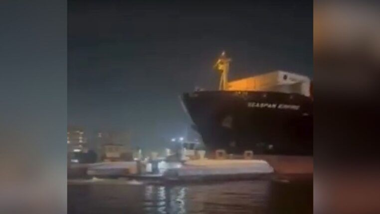 Registro noturno de um grande navio cargueiro escuro posicionado muito próximo a uma balsa de transporte de passageiros em um canal. A iluminação é artificial, vinda da embarcação e das luzes da cidade ao fundo, com a água refletindo o brilho das luzes.