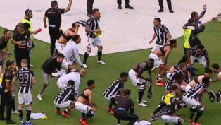 Jogadores e comissão técnica de um time de futebol, vestindo uniformes de listras verticais pretas e brancas com calções brancos, celebram intensamente em campo. Alguns atletas estão ajoelhados ou agachados no gramado, enquanto outros correm e agitam camisas no ar. Ao fundo, profissionais de imprensa com coletes e microfones acompanham a movimentação na borda do campo.